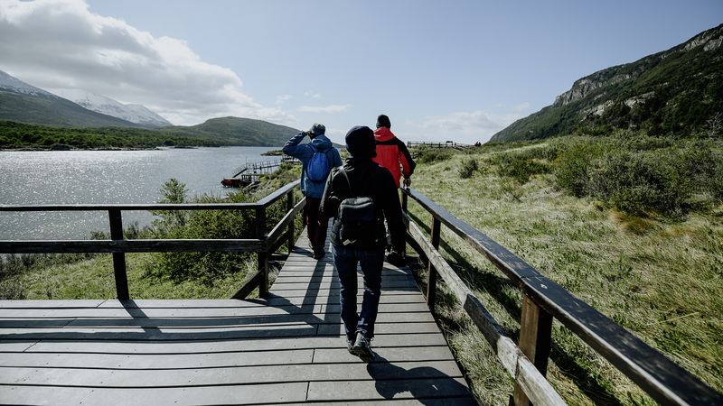Hikers on a wooden boardwalk overlooking a pristine lake surrounded by mountains in Ushuaia National Park, Patagonia.