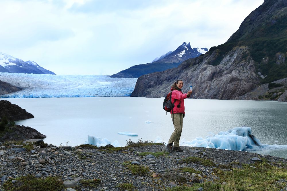 Hiker in red jacket photographs Glaciar Grey's blue ice chunks and calving face in Torres del Paine National Park, Patagonia.