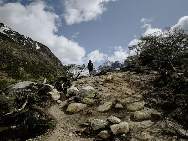 Hiker standing on rocky mountain trail with snow-capped peaks and twisted trees in Laguna Esmeralda trek, Patagonia.