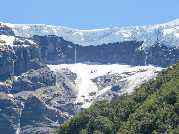 Glaciar Tronador descends between towering cliffs with cascading waterfalls, surrounded by green forest under clear blue skies.