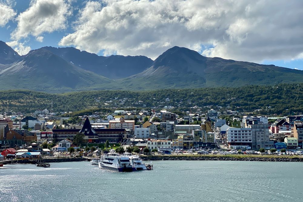 Ushuaia, Argentina: Waterfront city with boats, backed by forested hills and rugged mountains under a partly cloudy sky.