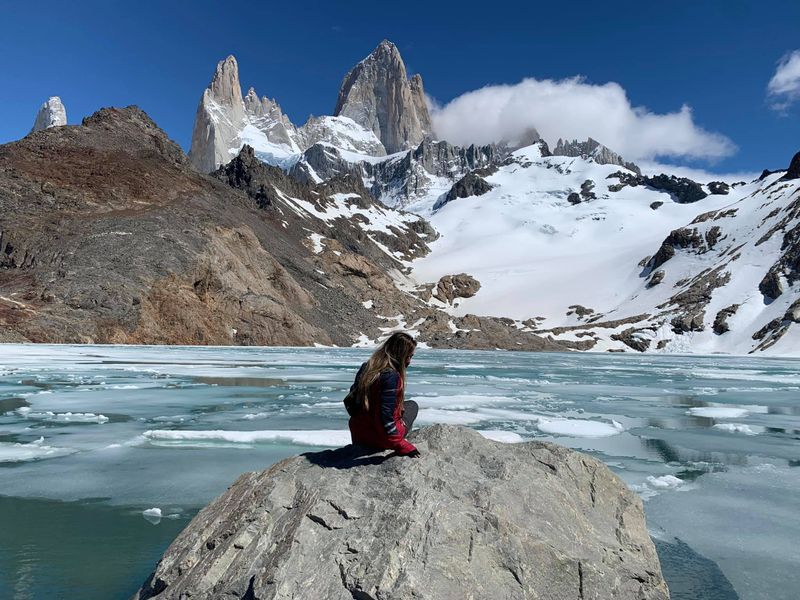 Woman sitting on rock overlooking turquoise glacial lake with snow-capped granite peaks of Mount Fitz Roy at Laguna de los Tres, Patagonia.