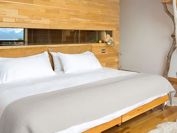 Modern bedroom with wooden architecture, white bedding, and panoramic window overlooking Patagonian landscape at Tierra Patagonia.