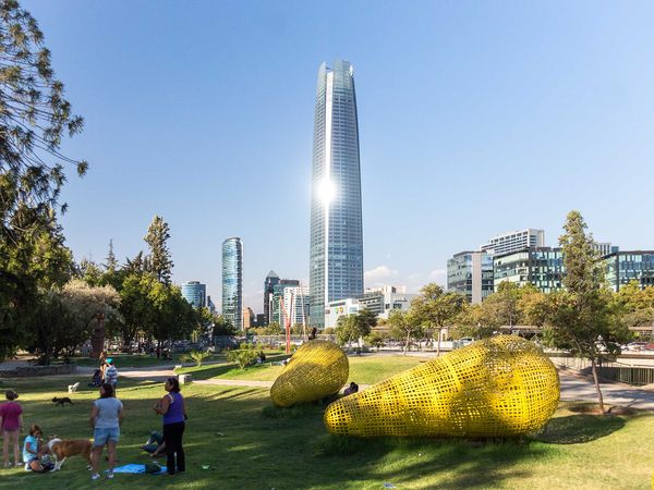 Modern Santiago skyline with tall contemporary tower overlooking a vibrant urban park featuring yellow sculptural installations and visitors enjoying green spaces.