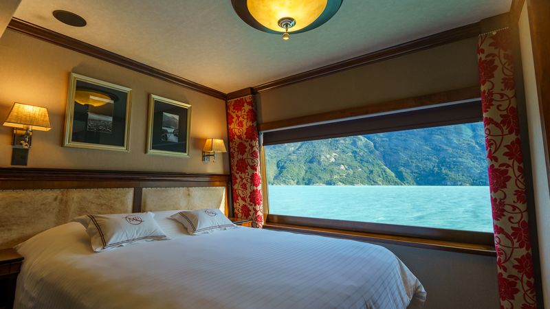 Luxury cruise cabin with panoramic window overlooking turquoise waters and forested mountains near El Calafate, Patagonia.