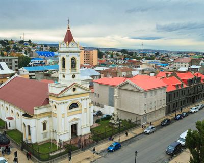 White church with red roof in Punta Arenas, Patagonia, surrounded by colorful buildings and mountains.