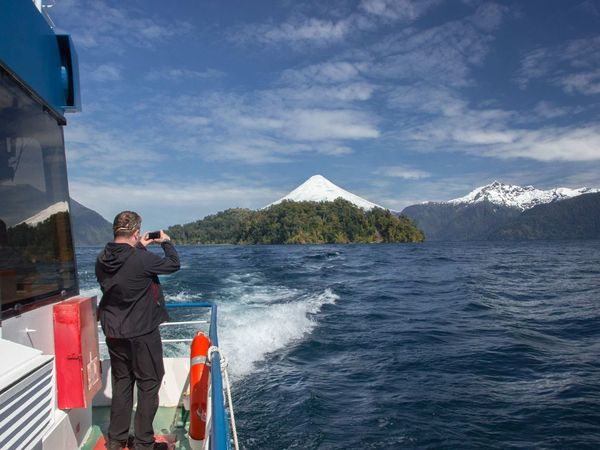 Tourist photographing snow-capped volcano and forested island from ferry boat on Lake Todos los Santos during Cruce Andino crossing near Puerto Varas.