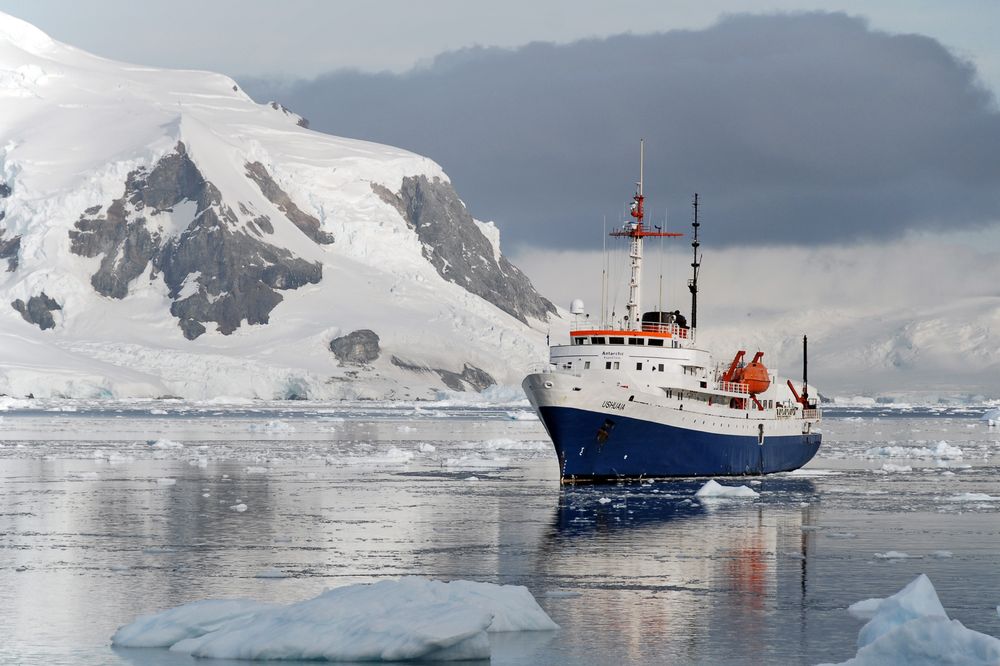 Research vessel navigates icy Antarctic waters near snow-capped mountain and glaciers.