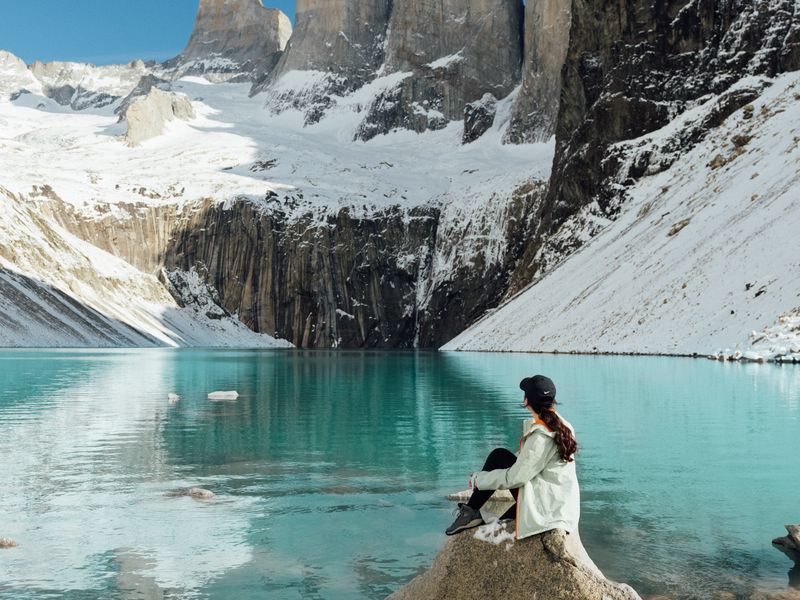 Woman sitting on rock overlooking turquoise glacial lake with snow-capped granite peaks of Torres del Paine, Patagonia.