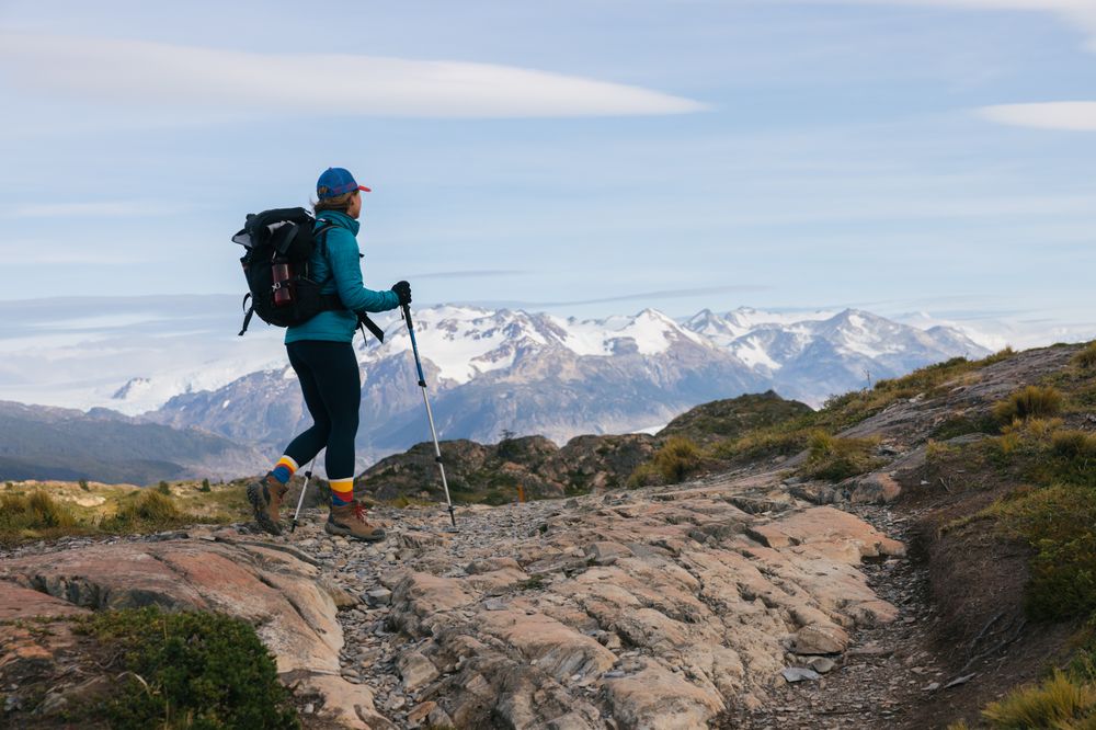 Hiker with backpack and trekking poles on rocky trail with snow-capped mountains in Torres del Paine, Patagonia.