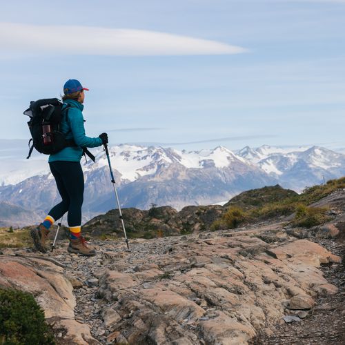 Hiker with backpack and trekking poles on rocky trail with snow-capped mountains in Torres del Paine, Patagonia.