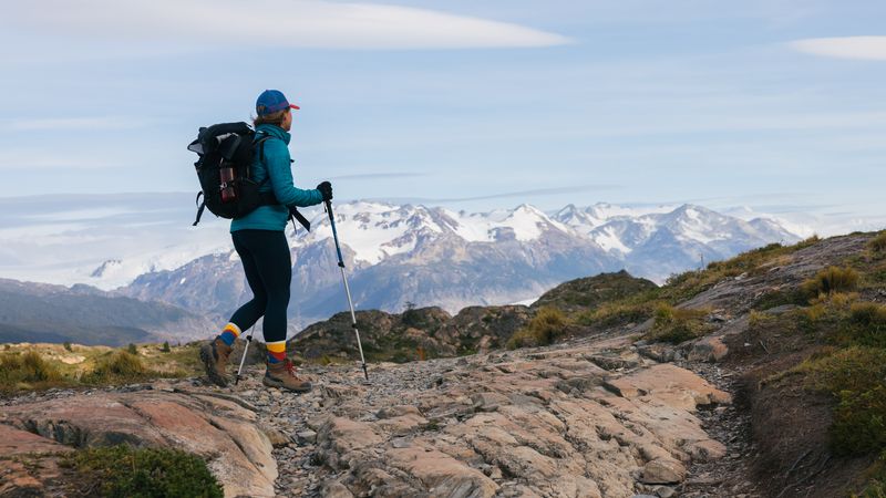 Hiker with backpack and trekking poles on rocky trail with snow-capped mountains in Torres del Paine, Patagonia.