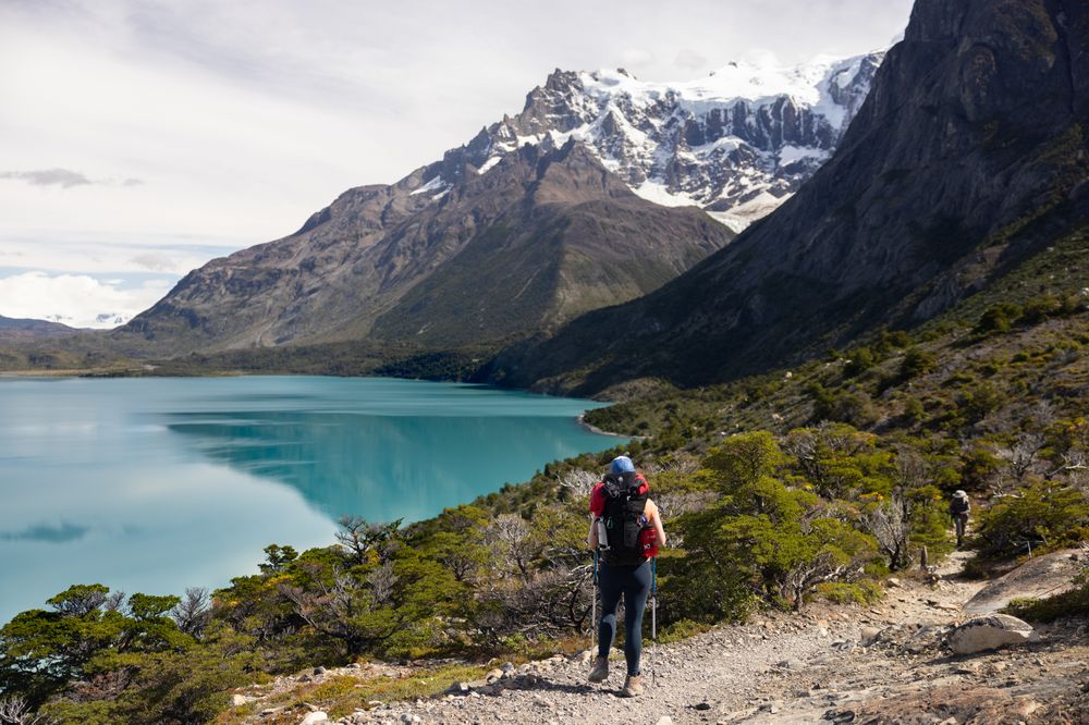 Hikers trekking along a turquoise glacial lake with snow-capped peaks of Torres del Paine rising dramatically in the background.