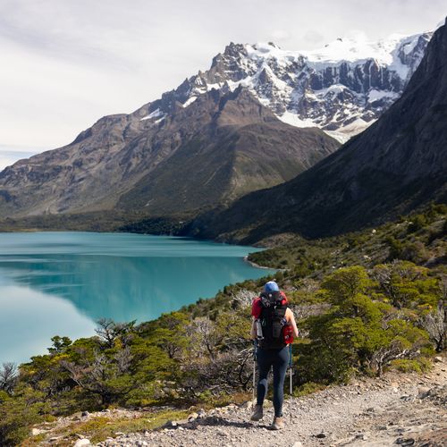 Hikers trekking along a turquoise glacial lake with snow-capped peaks of Torres del Paine rising dramatically in the background.