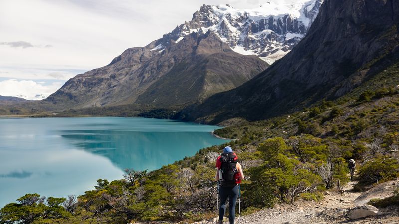 Hikers trekking along a turquoise glacial lake with snow-capped peaks of Torres del Paine rising dramatically in the background.