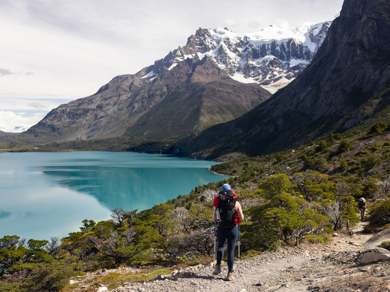Hikers trekking along a turquoise glacial lake with snow-capped peaks of Torres del Paine rising dramatically in the background.