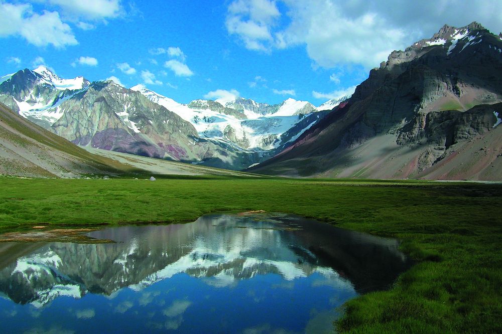 Alpine valley near Aconcagua with snow-capped peaks, glaciers, and mirror-like pond reflecting mountains under blue sky.