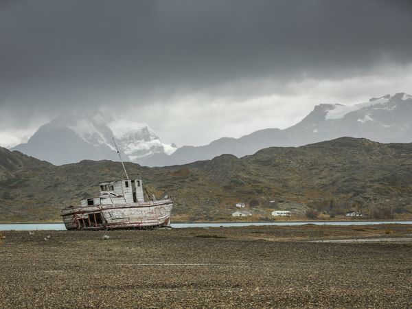Abandoned fishing boat beached on gray shale with snow-capped mountains under stormy skies in Patagonia.