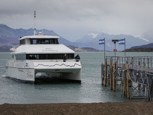 White catamaran Estancia Cristina docked at pier with snow-capped Patagonian mountains and glacier in background.
