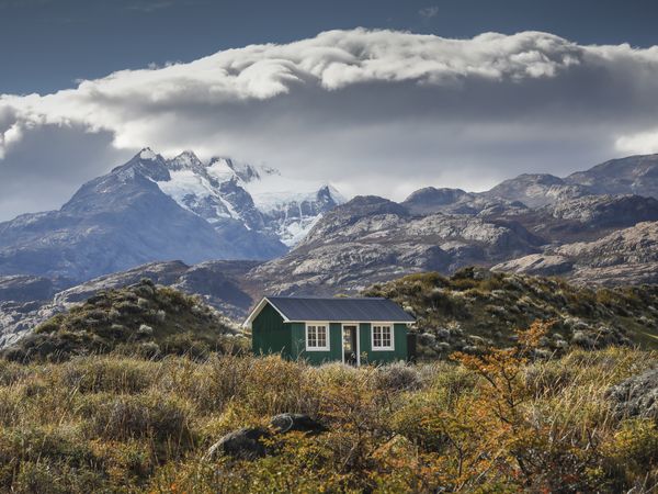 Green cottage nestled in windswept Patagonian steppe with snow-capped Andes mountains and dramatic clouds in background.