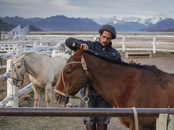 Gaucho prepares brown horse at Estancia Cristina ranch with snow-capped Patagonian mountains in background.