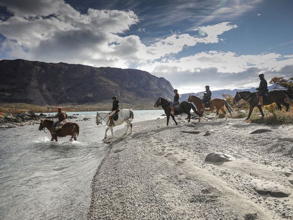 Group of horseback riders crossing a river with mountains and glaciers in Patagonia's Estancia Cristina region.