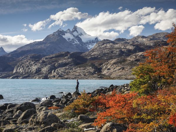 Hiker stands on rocky shore of turquoise lake with snow-capped mountain peak and autumn foliage at Estancia Cristina, Patagonia.