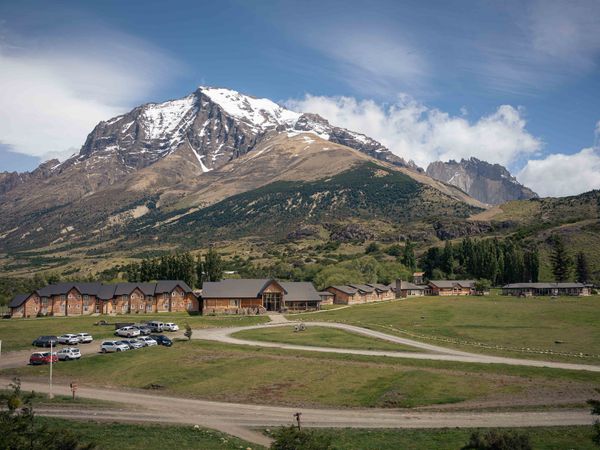 Wooden lodge buildings nestled in a green valley beneath snow-capped mountain peaks in Patagonia.