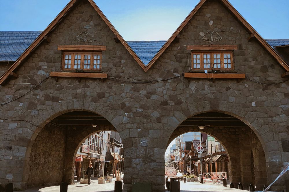 Twin stone archways frame a Western-style street in Bariloche, Argentina, under clear blue skies.