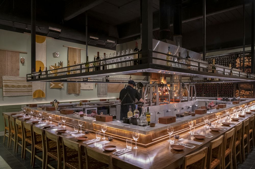 Modern Argentine asado restaurant in Buenos Aires featuring an open kitchen with chef, long communal dining counter, and traditional wood-fired grill setup.