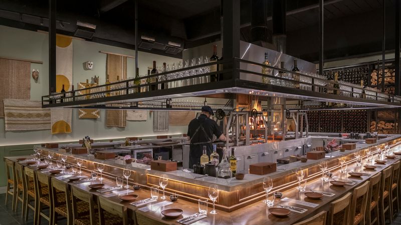 Modern Argentine asado restaurant in Buenos Aires featuring an open kitchen with chef, long communal dining counter, and traditional wood-fired grill setup.