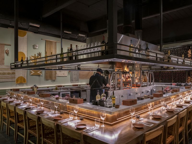 Modern Argentine asado restaurant in Buenos Aires featuring an open kitchen with chef, long communal dining counter, and traditional wood-fired grill setup.