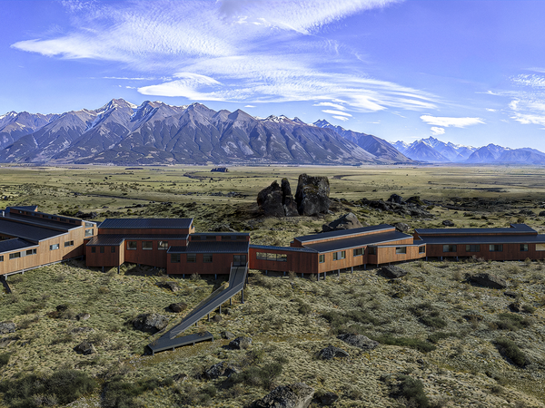 Modern lodge buildings nestled in vast Patagonian steppe with dramatic mountain range under blue sky.