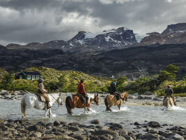 Horseback riders ford a river with snow-capped mountains and a traditional lodge at Estancia Cristina in Patagonia.