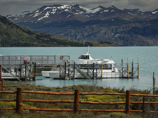 Boats docked at a turquoise glacial lake with snow-capped Patagonian mountains and green valleys in the background.