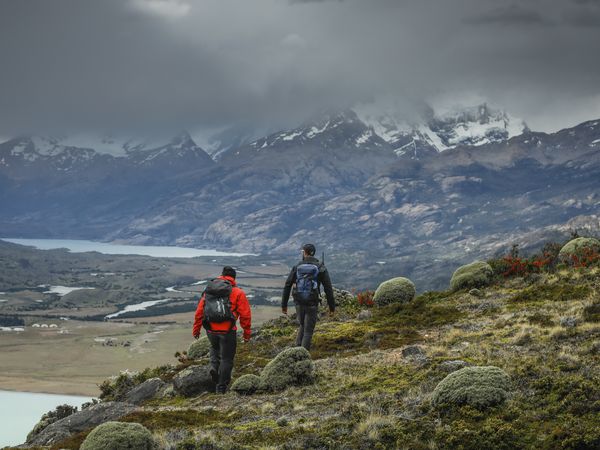 Two hikers trek through Patagonian wilderness toward snow-capped peaks and turquoise lakes near Estancia Cristina.