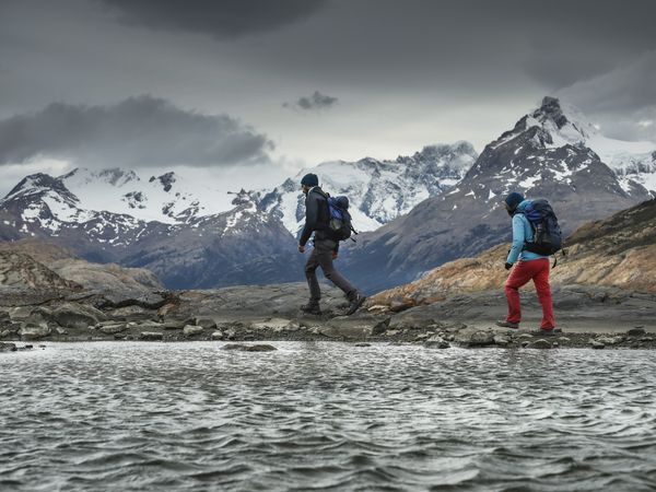 Two hikers trek across rocky terrain toward snow-capped peaks at Estancia Cristina in Patagonia.
