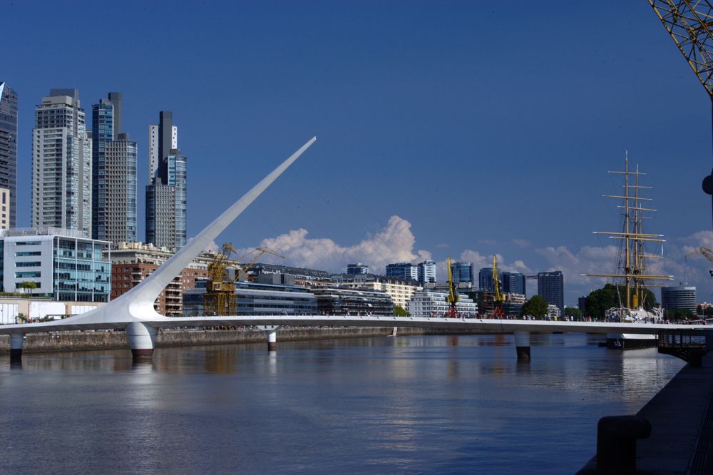 Buenos Aires Puerto Madero waterfront with iconic white bridge, modern skyline, and tall ship under blue sky.