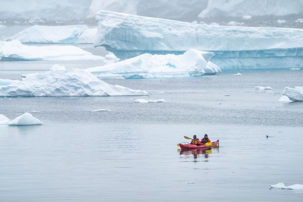 Two kayakers in a red boat navigate icy Antarctic waters surrounded by floating icebergs and glaciers.