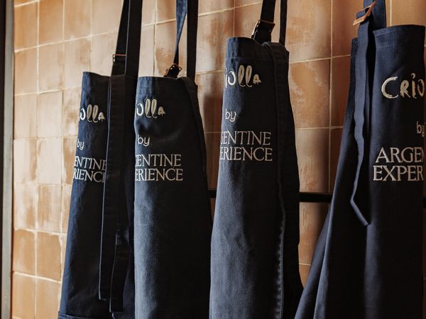 Dark denim aprons with "Argentine Experience" text hang on hooks against terracotta tiles in Buenos Aires.
