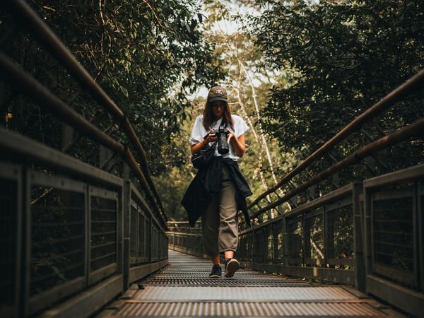 A woman in a cap walks on a metal bridge through a lush forest, looking at her camera.