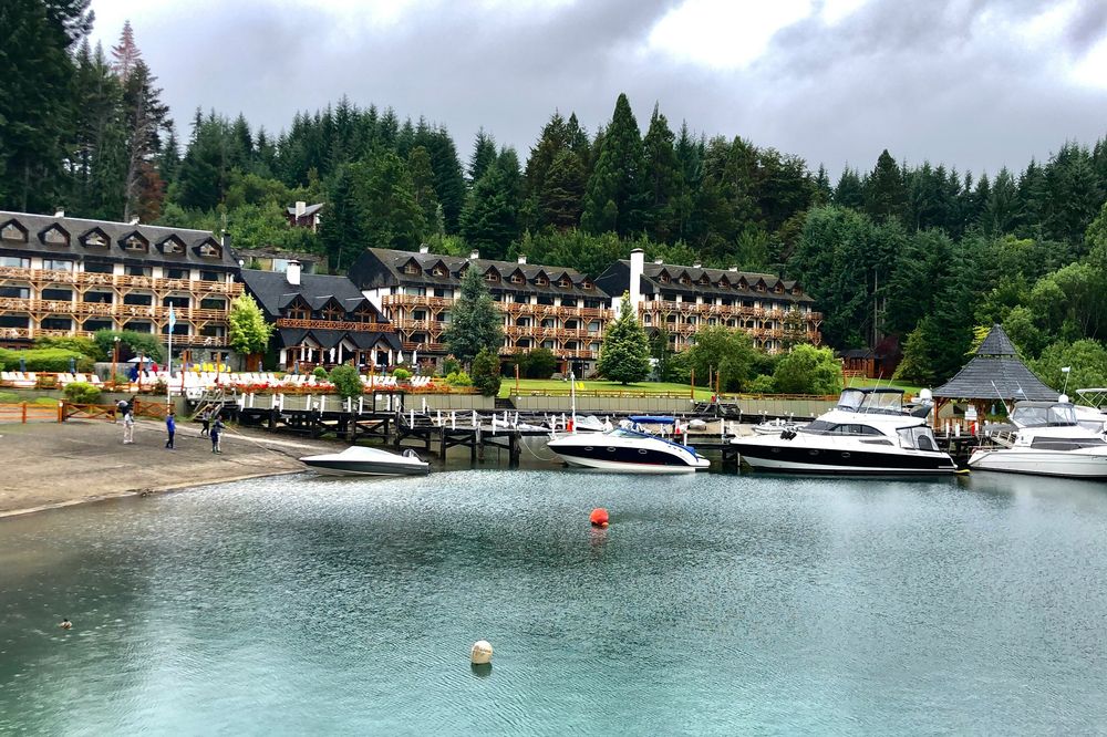 Lakeside lodge resort with wooden buildings, boats moored at dock, and forested mountains in Villa La Angostura.