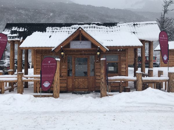 Snow-covered wooden lodge Los Cauquenes with mountain backdrop in Patagonia during winter snowfall.