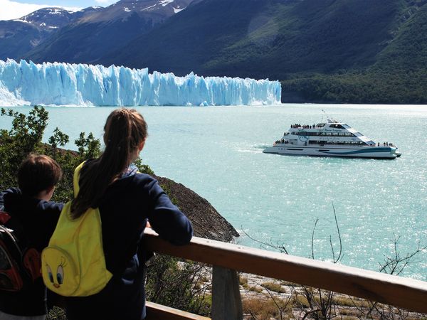Tourists viewing Glaciar Perito Moreno from a lookout platform while a tour boat floats on turquoise glacial lake waters in Patagonia.