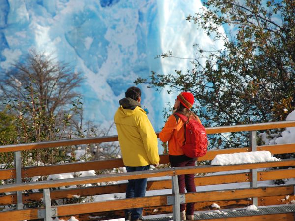 Two visitors in colorful jackets stand on wooden boardwalk viewing the blue ice of Glaciar Perito Moreno in El Calafate, Patagonia.