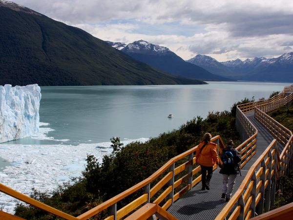 Wooden boardwalk overlooking turquoise glacial lake with snow-capped mountains and Perito Moreno Glacier in El Calafate, Patagonia.