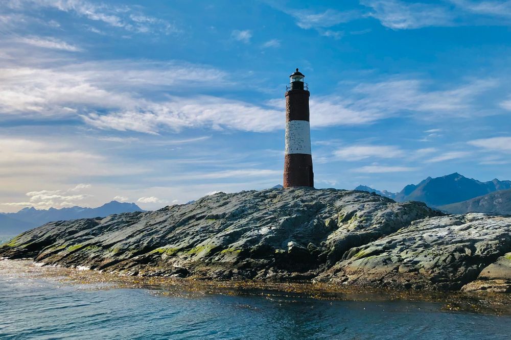 Red and white striped Faro del Fin del Mundo lighthouse stands on moss-covered rocky island in Beagle Channel near Ushuaia, Patagonia.