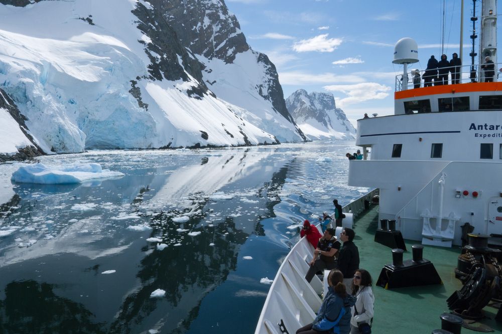 Expedition ship navigates icy Antarctic waters with glaciers and snow-covered peaks in the distance.