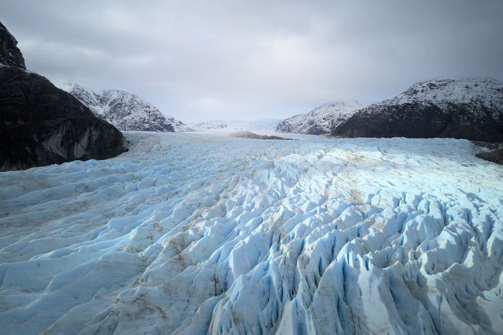 Vista aérea del Glaciar Guilliard fluyendo entre montañas nevadas en el fiordo de la Ruta Kaweskar, Patagonia chilena, bajo un cielo nublado