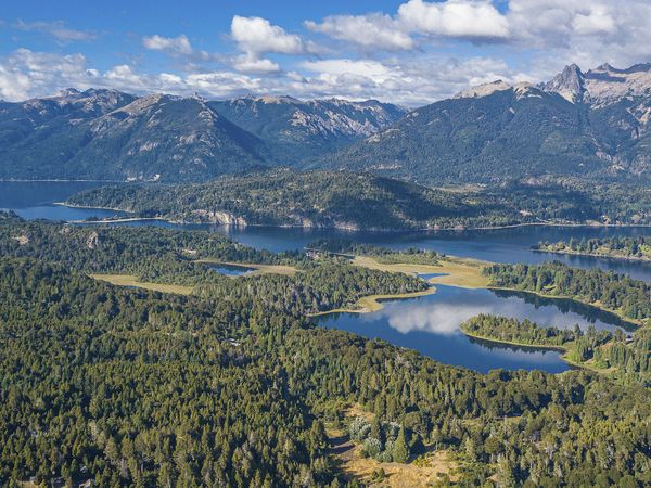 Aerial view of Bariloche's Circuito Chico featuring pristine alpine lakes, dense forest, and snow-capped Andes mountains under blue sky.